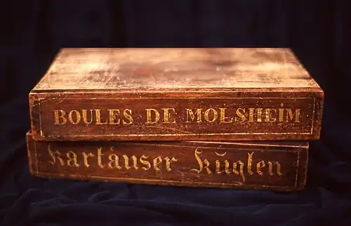 Deux boîtes de bois superposées, sur lesquelles sont écrits respectivement « Boules de Molsheim » et « Kartäuser Küglen.