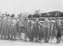 Photo en noir et blanc figurant des hommes portant sur leurs épaules un tronc passé au travers d'une monnaie de pierre