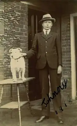 Photographie en noir et blanc d'un garçon debout, portant un costume et un chapeau, à côté d'un chien posé sur une table.