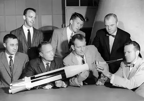 Sept hommes, quatre assis et trois debout, regardent une maquette de fusée.