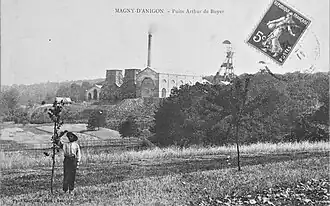 Photo en noir et blanc d'un ensemble de bâtiments industriels typés 1900 avec deux grands chevalements (tour avec bigue) métalliques et cheminée d'usine.
