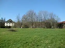 Un grand terrain couvert d'herbe, au fond se trouve un rideau d'arbre et les bâtiments d'une ancienne usine ainsi qu'une grande maison ancienne.