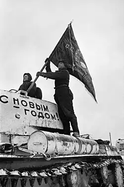 Photo en noir et blanc représentant un chef de char tête sortie de son tank, tandis qu'un soldat y hisse un drapeau. « Bonne année ! » est inscrit sur le char.