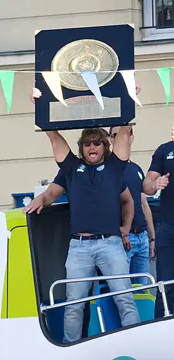 Homme en tenue de ville, en pied, tenant au dessus une planche représentant le bouclier de Brennus.