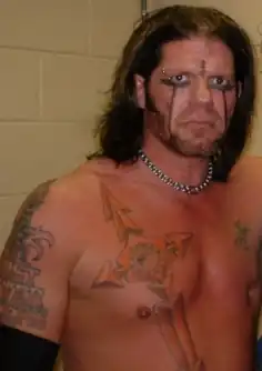 Photo d'un catcheur à hauteur du buste. Il se tient debout dans les coulisses. Il est torse nu, a les cheveux longs châtains foncés, porte un collier tour de cou en chaîne, a un maquillage imitant des coulées de larmes et arbore plusieurs tatouages sur les bras et un sur le torse en forme de rose des vents.