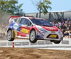 Photographie d'une voiture de rallycross bleue, blanche et rouge, en plein vol, après un saut, de trois-quarts.