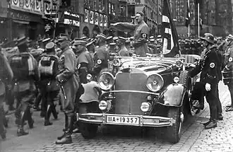 Photo noir et blanc prise en septembre 1935. Dans une rue de Nuremberg, Adolf Hitler, debout à l'arrière d'une voiture au premier plan, salue des troupes de miliciens de la SA défilant sur sa gauche. On distingue aussi, à l'arrière de la voiture, juste derrière Hitler, le drapeau à croix gammée de cérémonie, Blutfahne, et, en uniforme SS, debout à droite, devant la portière de la voiture, Jakob Grimminger, porteur officiel du Blutfahne.