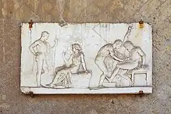 bas-relief, à gauche, Hercule nu debout devant une femme assise ; à droite, un soignant penché au-dessus de la cuisse d'un blessé assis