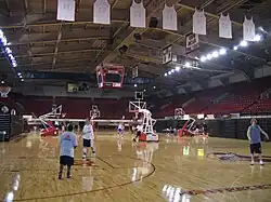 Vue de l'intérieur d'une salle de basket-ball dans laquelle les sièges des tribunes sont rouges. Des maillots blancs sont accrochés au plafond.