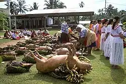 Photo en couleur montrant des cochons tournés les pattes en l'air, posés sur le sol d'une grande pelouse, à côté de paniers en osiers remplis de légumes et recouverts de nattes. Des gens sont présents à côté.