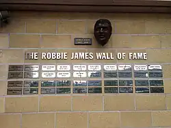 Photo d'un mur de briques orné d'une série de plaques en métal portant des noms et des dates. Elles sont surplombées par une inscription en grandes lettres blanches indiquant « The Robbie James Wall of Fame » et d'un buste en bronze représentant un homme moustachu aux cheveux courts.