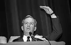 Photographie en noir et blanc d'un homme en costume et faisant un discours devant des micros