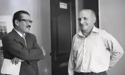 Un homme brun et moustachu en costume, portant des lunettes, et tenant un dossier dans la main discute avec un européen chauve d'un certain âge, vêtu d'une chemise blanche à manche longue.