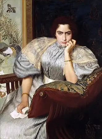 Portrait d’une femme assise dans un fauteuil en velours rouge, appuyée sur l’accoudoir avec une main soutenant son menton. Elle porte une robe rayée aux manches bouffantes ornées de dentelle, un bracelet doré et un collier décoré. Son regard intense et expressif est dirigé vers le spectateur. En arrière-plan, un mur décoré de motifs floraux et un oiseau peint ajoutent une touche de raffinement à la scène.