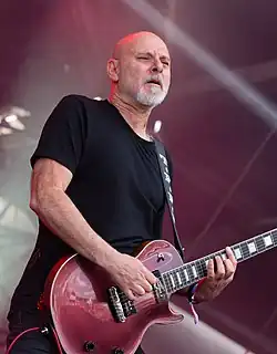 Photo d'un homme chauve avec une petite barbiche blanche. Il porte un tee-shirt noir et joue de la guitare électrique sur une scène.