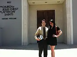 Man with light skin and dark hair and woman with dark hair and light skin stand in front of a building with a sign that reads The Church of Jesus Christ of Latter-day Saints; Visitors Welcome