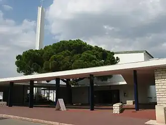 Photographie du temple formé d'un bâtiment blanc rectangulaire percé d'une grande porte, à demi caché par un pin parasol. L'arbre pousse dans une cour au revêtement rougeâtre, entourée d'une colonnade faite de minces piliers bleu vif supportant un toit de béton plat. Des murs et murets recouverts de pierres blanches rectangulaires complètent l'ensemble de l'architecture. À gauche se dresse un mince pilier de béton blanc supportant en haut une croix et, vers la moitié, une couronne de cloches.