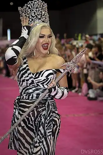 Photographie d'une femme trans blonde sur un défilé. Son visage exprime de la joie tandis qu'elle pose en tenant sa grande couronne de strass contre la tête d'une main et un sceptre de l'autre.