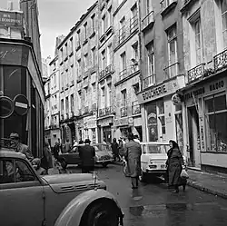 Photo en noir et blanc d'une rue entourée de modestes immeubles à trois étages aux façades défraîchies. Les enseignes des commerces sont en français et en hébreu, et arborent des étoiles de David. Quatre voitures et quelques passants apparaissent.