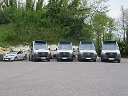 Photographie en couleurs de cinq véhicules garés côte-à-côte, vus de face : une voiture électrique blanche et quatre minibus bleu, blanc et noir. Il sont stationnés devant un mur en pierre blanches et des arbres.