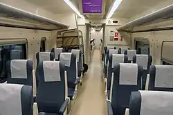 Photo couleur montrant un intérieur de train de couleur crème, sol, parois et plafond et sièges individuels bleu à têtière blanche.