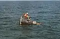 Photographie couleur d'une capsule Gemini dans l'eau avec les deux astronautes et un plongeur visibles.