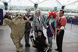 Groupe de six personnes portant des costumes. Celui de droite est une personnification d'Ed Wood, avec une petite moustache et un pull rose porté par-dessus un costume.