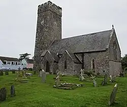 Photo d'un bâtiment allongé aux murs de pierre grise et au toit en ardoises avec une grande tour carrée crénelée munie d'une horloge
