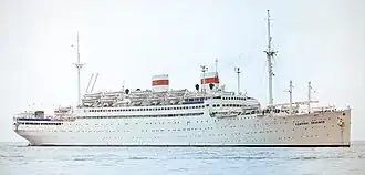 Photographie en couleurs d'un gros navire blanc avec deux cheminées. Celles-ci sont ceinturés par une ligne rouge et le logo de l'URSS. Il est vu sur une mer calme.