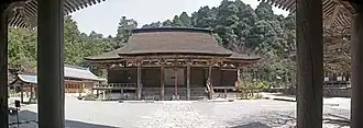 Photo couleur de l'enceinte d'un sanctuaire (sol pierreux et lumineux de couleur blanchâtre). On distingue un pavillon d'ablution, le bâtiment principal en bois doté d'un toit de chaume et une pagode à trois étages. À l'arrière-plan, une forêt d'arbres verts sous un ciel blanc laiteux.