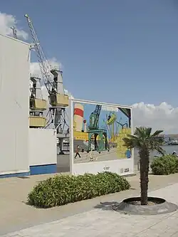 Photographie d'une place avec des installations portuaires à l'arrière-plan.