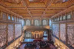 Photo de l'intérieur d'une immense salle. Les murs de droite et de gauche portent d'immenses fresques. Le mur de face porte une fontaine et des baies vitrées.