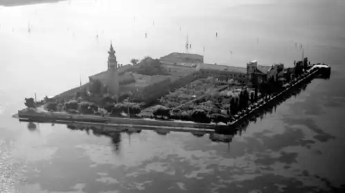 Photographie en noir et blanc prise depuis un avion. On voit une île rectangulaire occupée par de nombreux bâtiments contigus, entourés de jardin.