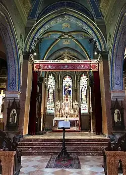 Photographie couleur du chœur de l'église, séparé de la nef par quatre marches. Le plafond est peint en bleu avec des motifs dorés. Au centre l'autel entouré de vitraux.