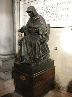 Statue sur un socle représentant un personnage assis, portant une toque et une capuche, tête baissée