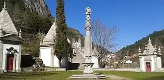 Photo d'une colonne de pierre surmontée d'une statue, avec quelques chapelles de part et d'autres, et au fond un grand escalier menant à une église à deux clochers