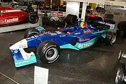 Photographie d'une monoplace de Formule 1 bleu foncé et vert clair, vue de trios-quarts, dans un musée.