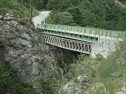Un pont en croisillons de bois franchit un ravin, remplacé par un autre pont à poutre métallique construit juste au-dessus.