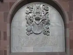 photographie en couleur montrant une niche creusée dans un mur en grès rose dont le fond en pierre grise est décoré par un relief avec des armoirie, en dessous desquelles se trouve une inscription.