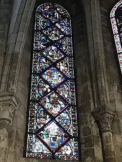 Vitrail composé de pièces rectangulaires et triangulaires. Sont représentés différents personnages dans plusieurs scénettes, difficilement visibles en raison du noircissement du vitrail.