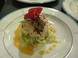 Au centre d'une assiette blanche à fin bord vert, un moulage de coquilles Saint-Jacques est posé en équilibre sur des morceaux de fenouil cuits et surmonté d'une fleur de couleur bordeaux et de quelques feuilles. De la sauce orange pâle entoure partiellement l'ensemble.