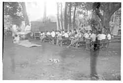 Photo en noir et blanc d'écoliers assis sur des bancs en plein air regardant un grand tableau.