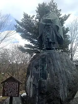 Photo couleur montrant, sur fond de ciel bleu nuageux, une statue en pied d'un homme en tenue de moine sur un rocher gravé d'une inscription.