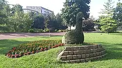 Au milieu d'un espace de pelouse verte, près d'un chemin piéton en asphalte rose, se dresse une sculpture végétale représentant un paon. Placée sur une petite butte maintenue par un mur de pierre de quelques marches, le corps de l'animal est constitué d'un seul arbuste taillé en forme de corps, cou et tête. Un cône métallique vient symboliser le bec. La queue de l'animal, posée au sol derrière lui, est composée d'un parterre de fleurs orange et rouges sur le pourtour.