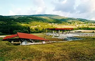 Vestiges avec colonnes et murs, partiellement recouvert par une toiture moderne de protection avec une colline, des champs et des bois en arrière-plan.