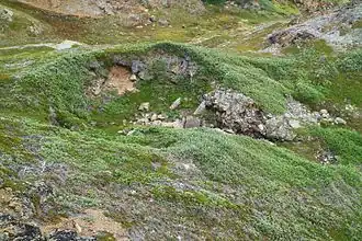 Photographie en couleurs des ruines d'une habitation en pierre situées dans le creux de roches affleurantes recouvertes de lichens.