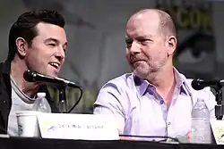 Photo de deux hommes assis derrière des tables et devant des microphones.