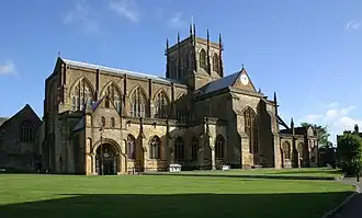 Photo d'un bâtiment allongé aux grandes fenêtres en ogive muni d'une grande tour carrée en son centre. Il se trouve au milieu d'un pelouse verte, avec quelques bâtiments visibles à l'arrière-plan.