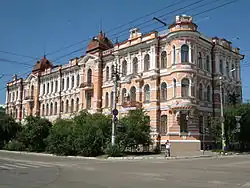 Bâtiment historique aux façades ornées de briques rouges et blanches, typique de l'architecture classique ou néo-Renaissance. Il est entouré d'arbustes et de rues dégagées sous un ciel bleu clair.