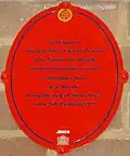 Plaque rouge ovale indiquant : « En mémoire du "District Officier" Charles Pearson de la "London Fire Brigade" qui est mort de ses blessures au numéro 100 Sidney Street, proche de ce site pendant le Siège de Sidney Street, le 3 janvier 1911 ».